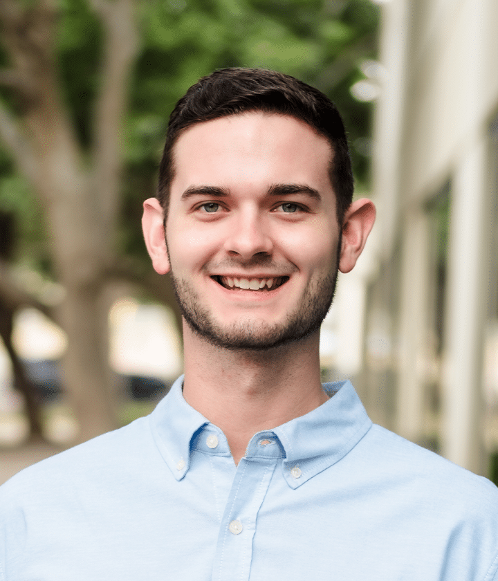 A young man with short dark hair and a trimmed beard smiles outdoors, wearing a light blue button-up shirt. Trees and a building are blurred in the background.