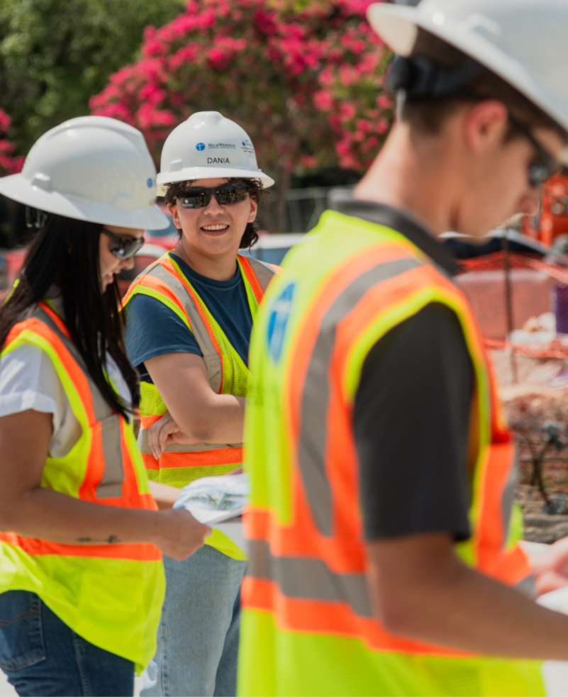 Three construction workers wearing safety vests and helmets stand outdoors at a work site, with one of them smiling.