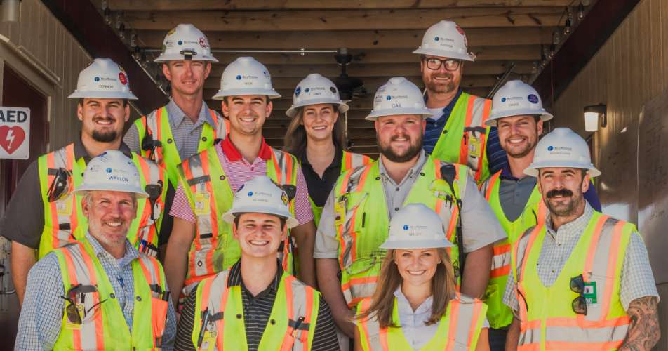 A group of construction workers in safety vests and hard hats pose for a group photo in a covered outdoor area.