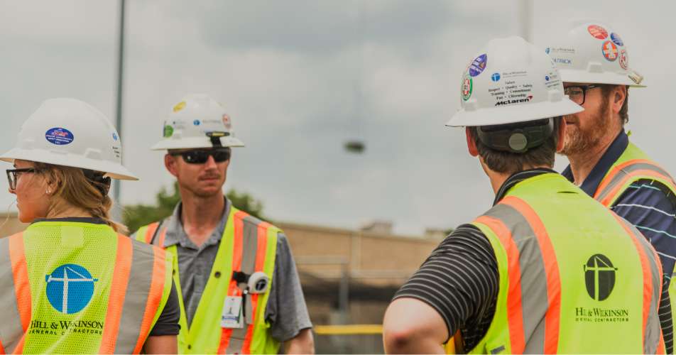 Four construction workers wearing safety vests and hard hats stand together at a job site, engaged in conversation.