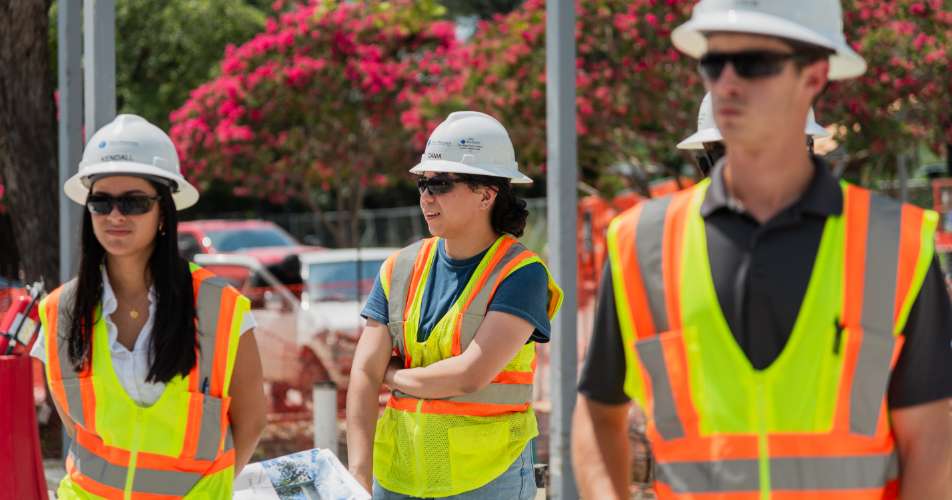 Three construction workers wearing safety vests, sunglasses, and hard hats stand outdoors near trees with pink flowers.
