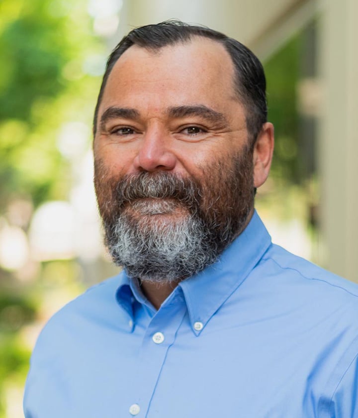 A man with a gray beard and short dark hair, wearing a light blue button-up shirt, stands outdoors in front of a blurred background with trees and buildings.