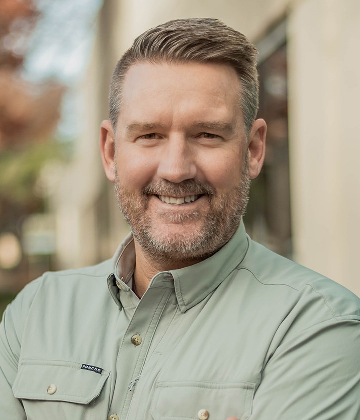 A man with short light brown hair and a trimmed beard smiles at the camera, wearing a light green button-up shirt, standing outdoors near a building.
