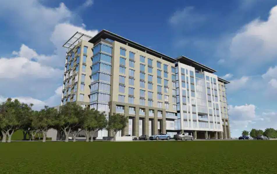 A modern multi-story office building with large windows, surrounded by trees and parked cars, under a blue sky with clouds.