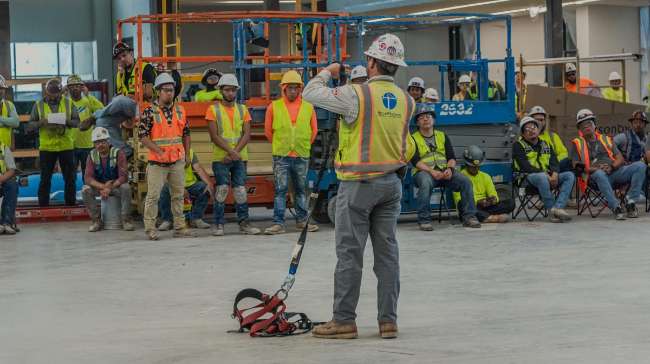 A construction safety trainer demonstrates equipment to a group of workers wearing safety vests and hard hats inside an unfinished building.