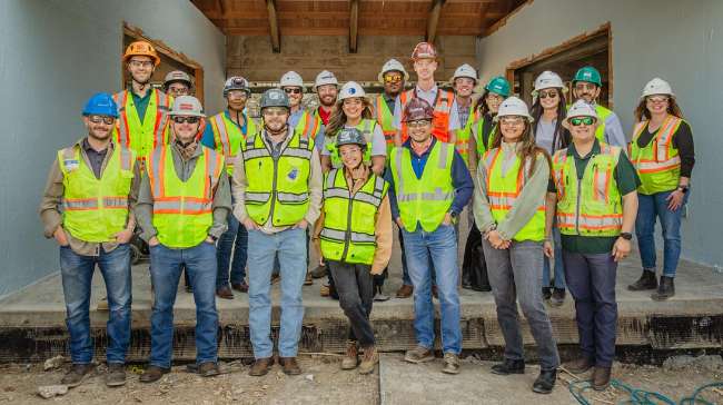A group of construction workers wearing safety vests, helmets, and work boots pose for a photo on a building site.