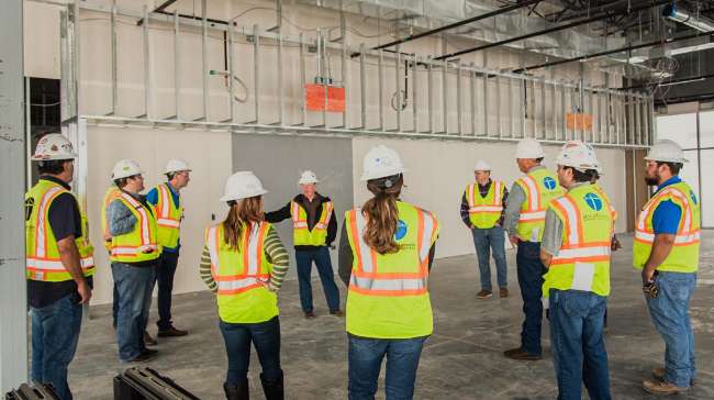 A group of construction workers wearing safety vests and hard hats stand in a semi-circle indoors, listening to a supervisor at a building site.