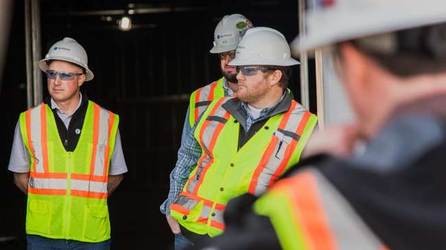 Four construction workers wearing safety vests, hard hats, and protective glasses stand together indoors at a job site.