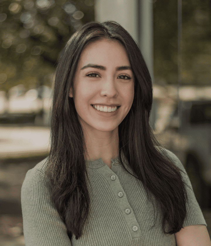 A woman with long dark hair, wearing a ribbed, button-up top, smiles while standing outdoors with trees and cars blurred in the background.