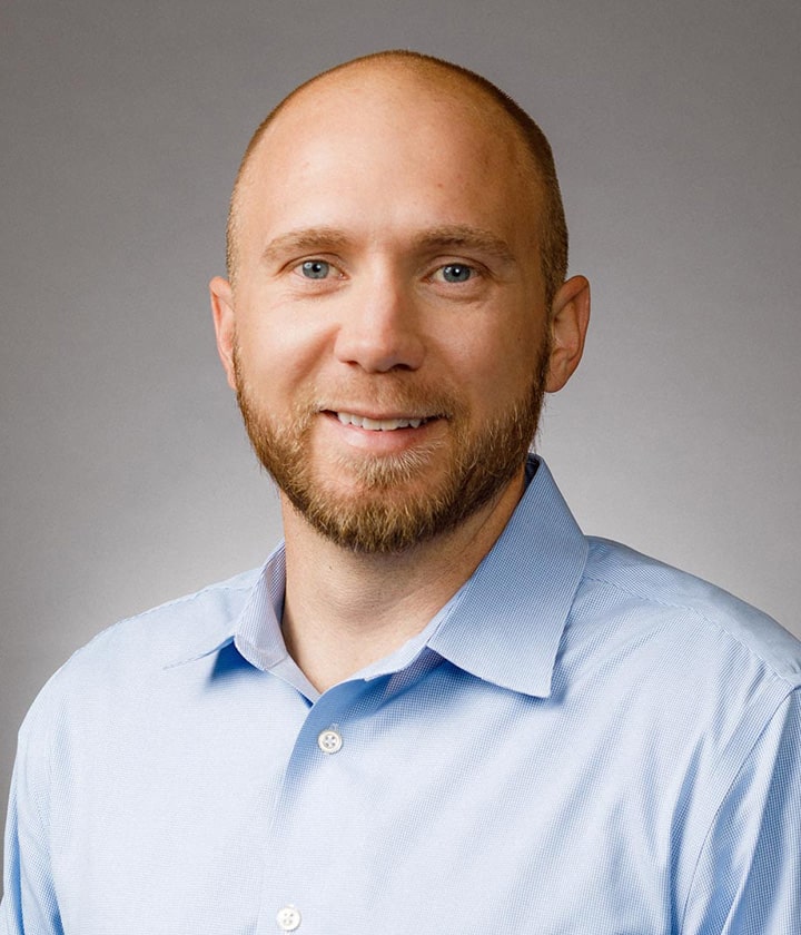 A man with a beard and short hair, wearing a light blue collared shirt, smiles at the camera against a plain gray background.
