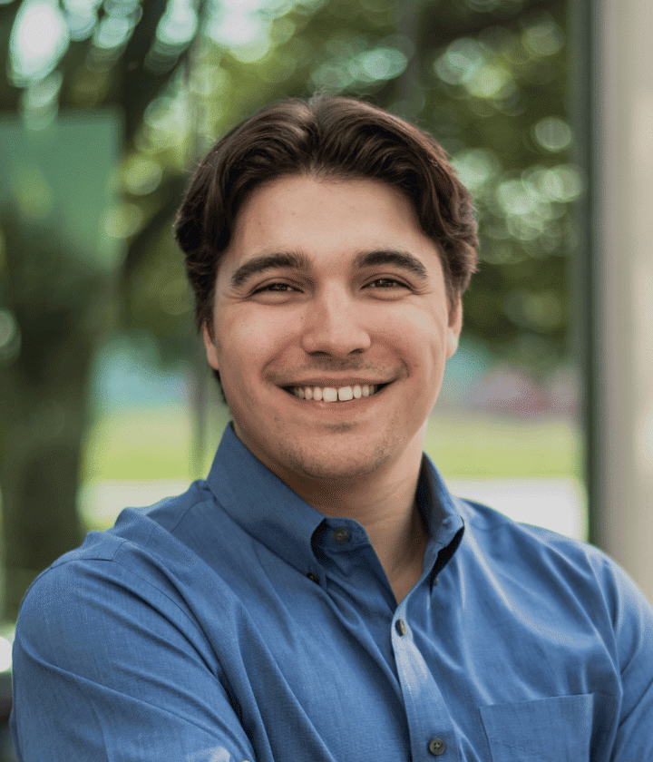 A man with dark hair and a blue button-up shirt smiles at the camera while standing in front of a window with greenery outside.