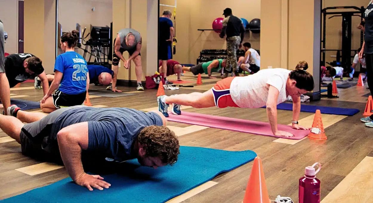 A group of people performs floor exercises on mats in a gym, including push-ups and planks, with cones and workout equipment visible around the room.