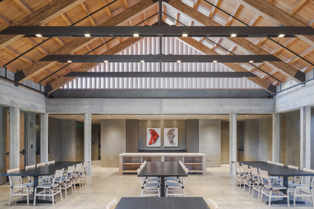 A photo of an open-floor plan dining area inside of a building with wood beams on the ceiling, and tables and chairs.