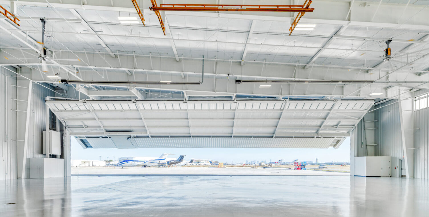 Inside of an airplane hangar with the large vertical-opening hangar door partially open to the outside, where a plane is sitting on the tarmac