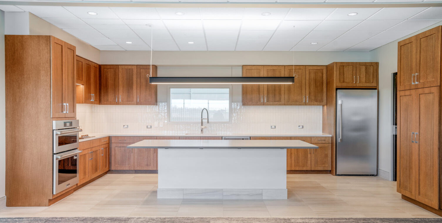 Interior of an office breakroom with brown cabinets, white countertops, an island in the middle, a sink, dishwasher, and refrigerator