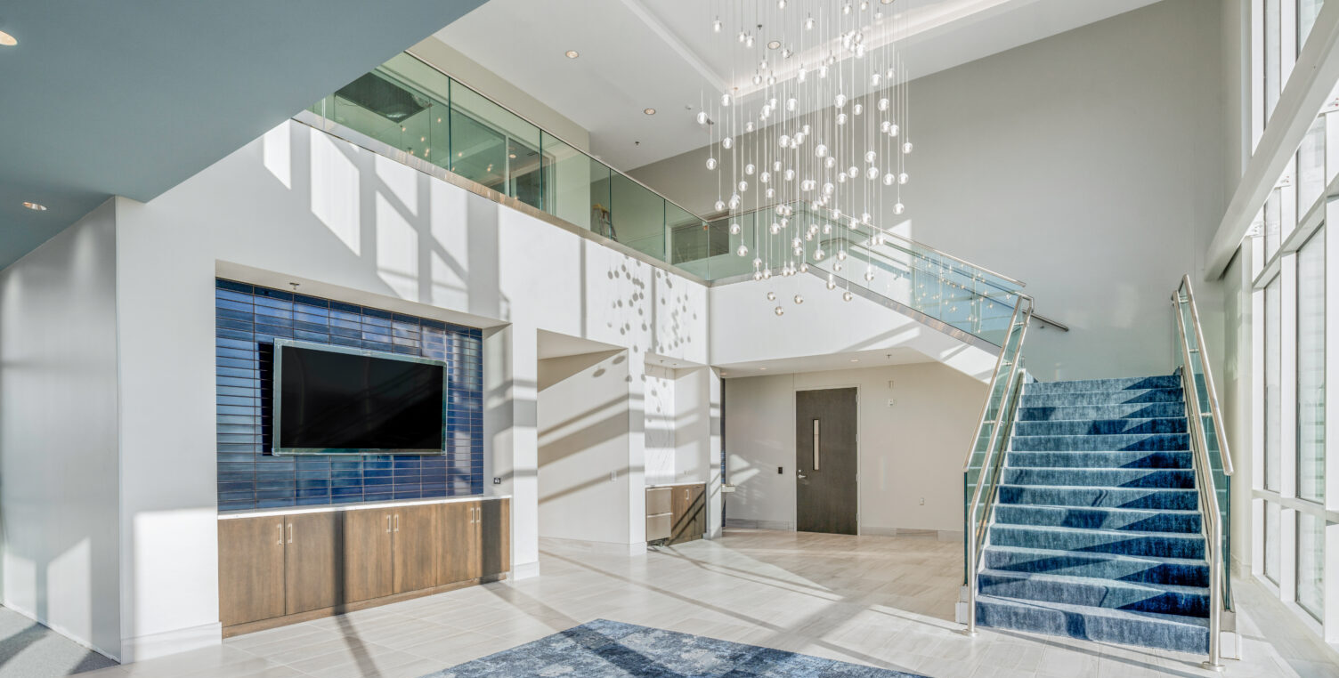 Lobby area of an office building with a large blue rug, stairs leading up to the second floor, a TV on the wall, a hanging light fixture in the middle of the room, and sunlight streams through the windows into the room from behind where the camera was positioned