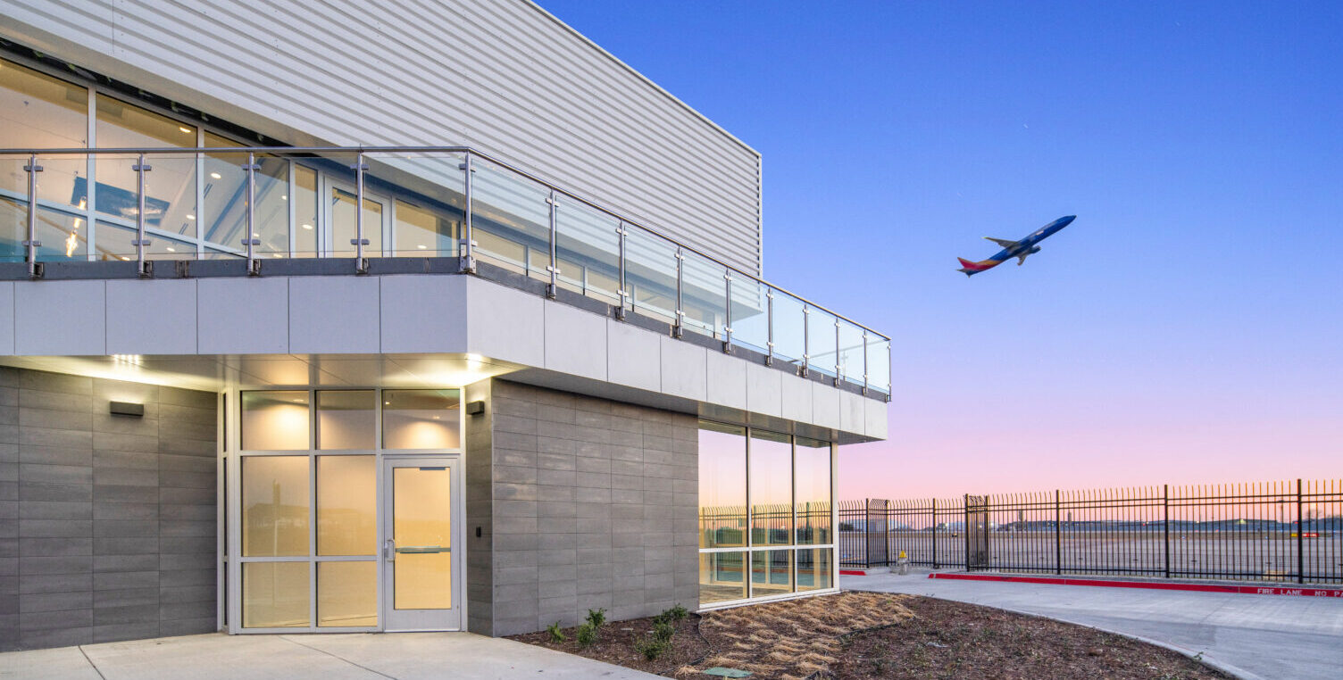 Exterior corner of an office building at early morning sunrise next to an airport runway with a plane at mid-take-off in the sky