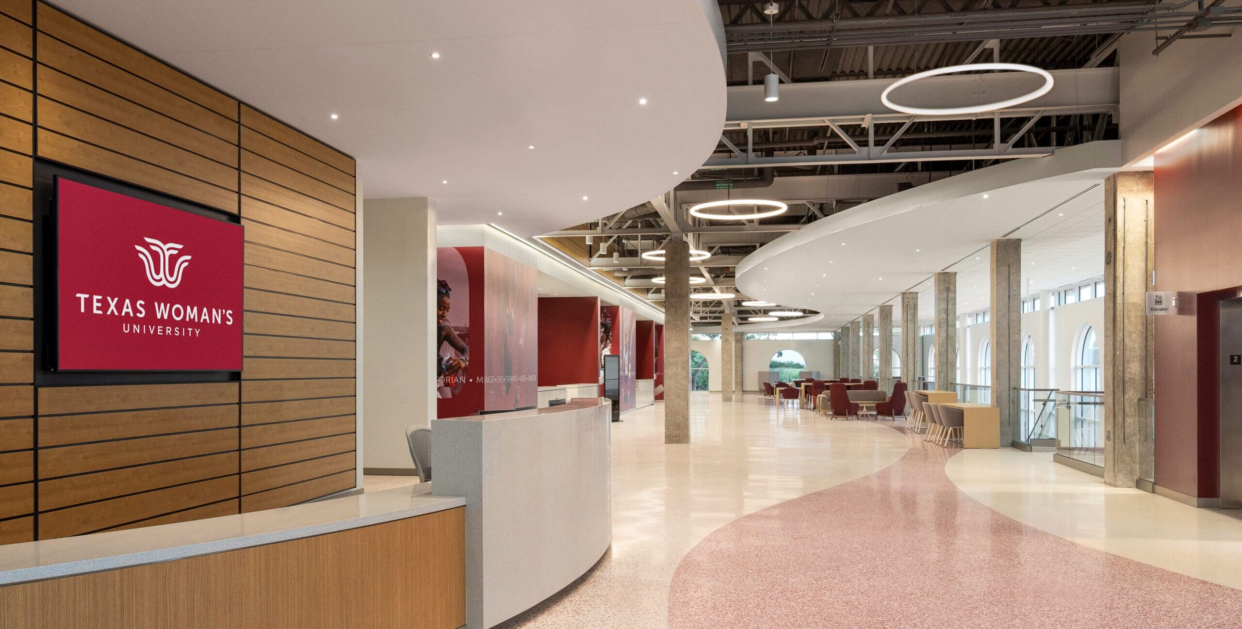 A modern interior lobby features a welcome desk and open seating area with upholstered lounge chairs, round tables, and collaborative workspaces. Exposed steel beams and circular pendant lighting hang from the high ceiling above polished flooring. Tall windows along one side bring in natural light, while red accent walls and glass-fronted office or reception spaces line the opposite side.