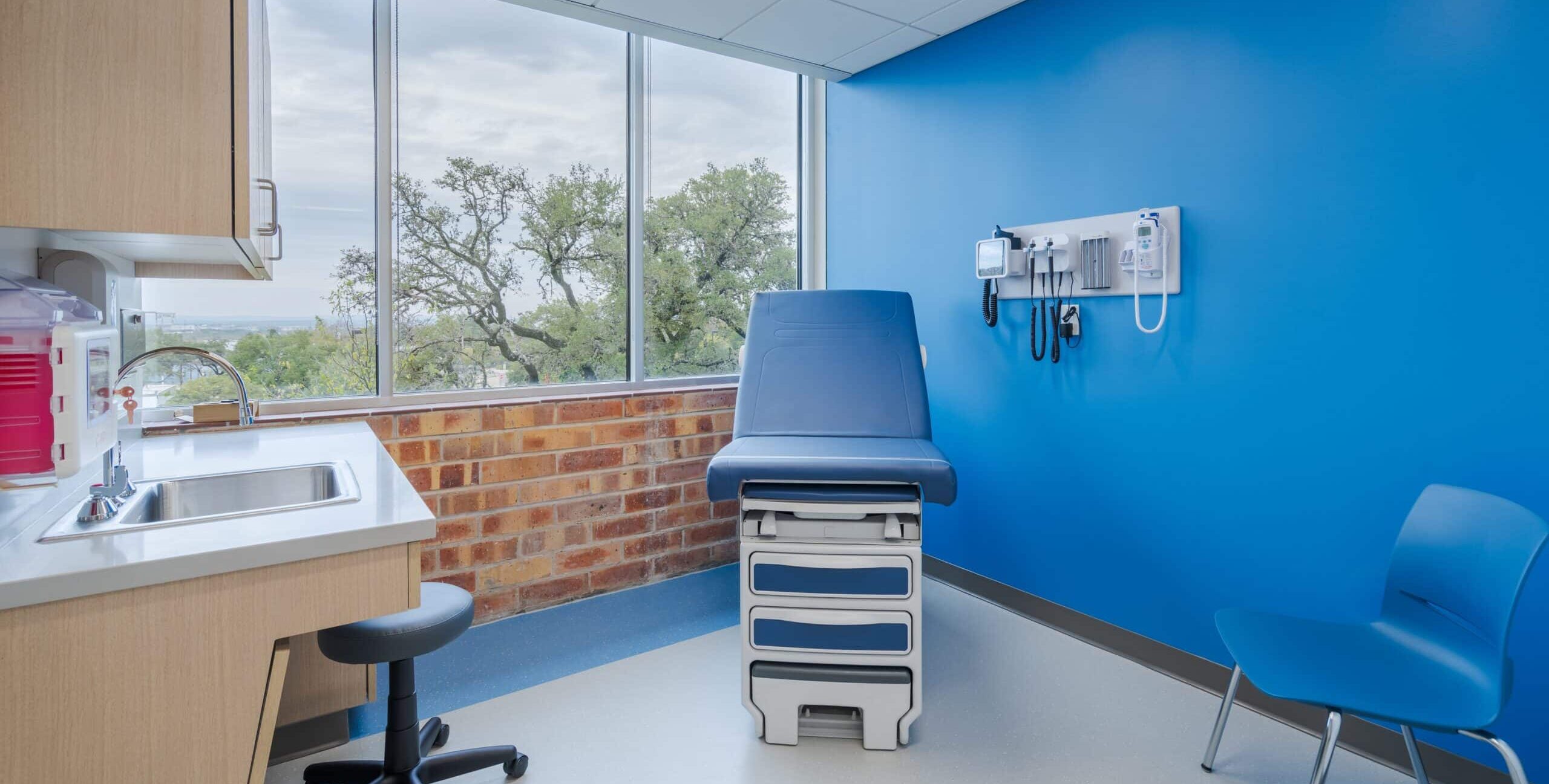 A clean medical examination room with a blue exam table near a large window showing trees outside. The walls are bright blue with a section of exposed brick under the window. A panel with medical instruments—such as a blood pressure monitor, otoscope, and thermometer—is mounted on the right wall. On the left side, there is a countertop with a sink, faucet, and wooden cabinets above, with a stool tucked underneath. A blue chair sits beside the exam table. The room is brightly lit and neatly organized.