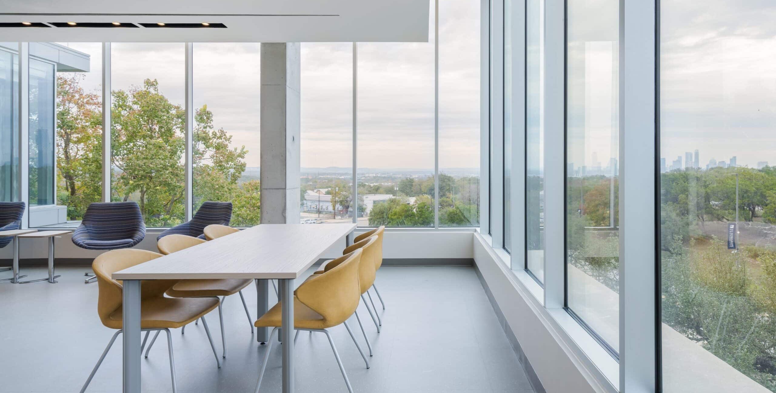 A modern indoor meeting space with large floor‑to‑ceiling windows overlooking trees, nearby buildings, and a distant city skyline. A long rectangular table with six mustard‑yellow chairs sits in the center of the room. Several blue lounge chairs are arranged near the windows. The space has light-colored floors and walls, a minimalist design, and abundant natural light.