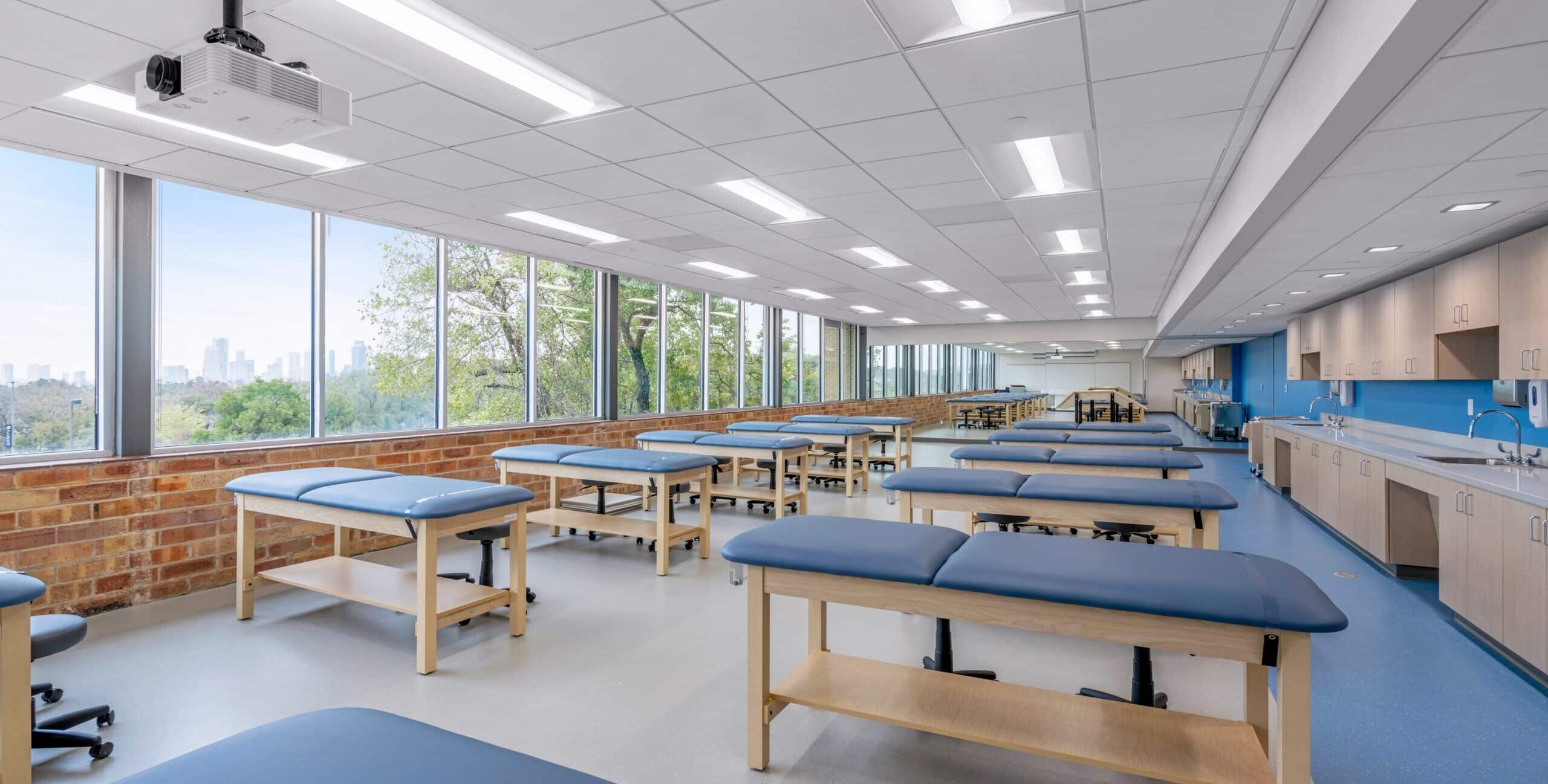 A large, bright classroom or training lab with multiple blue padded examination tables arranged in rows. One long wall is lined with windows showing trees and a distant city skyline. The room has recessed ceiling lights, a mounted projector, and a side wall with cabinets, sinks, and countertops. The space appears organized and designed for medical or physical therapy instruction.