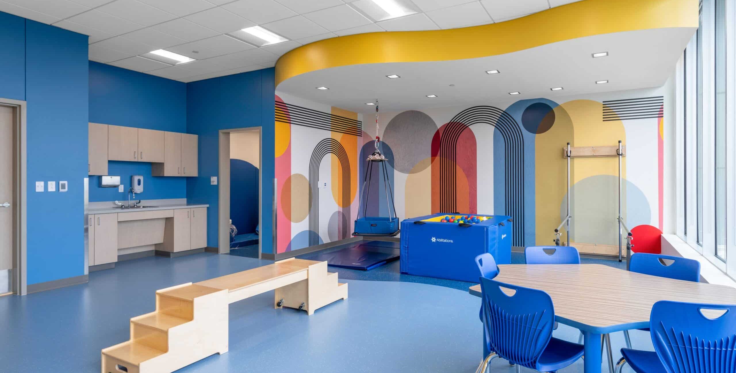 A brightly colored pediatric therapy room with blue floors and walls featuring a playful mural of geometric shapes in red, yellow, orange, gray, and blue. The room includes a wooden step structure, a blue padded therapy swing hanging from the ceiling, and a blue padded ball pit. A round table with blue chairs sits near built‑in cabinetry with a sink. Large windows let in natural light.