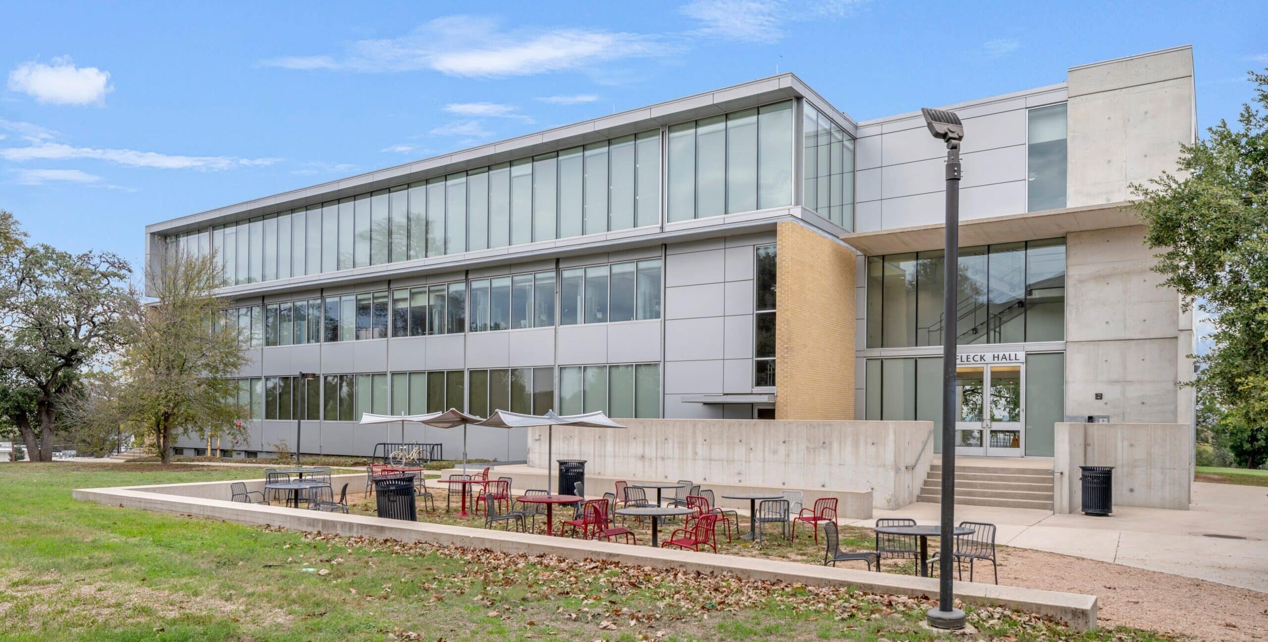A modern academic building with large glass windows, light metal panels, and sections of tan brick. In front of the building is an outdoor seating area with red and black tables and chairs, along with trash bins and a lamp post. The surrounding area includes grass, trees, and a blue sky with scattered clouds.