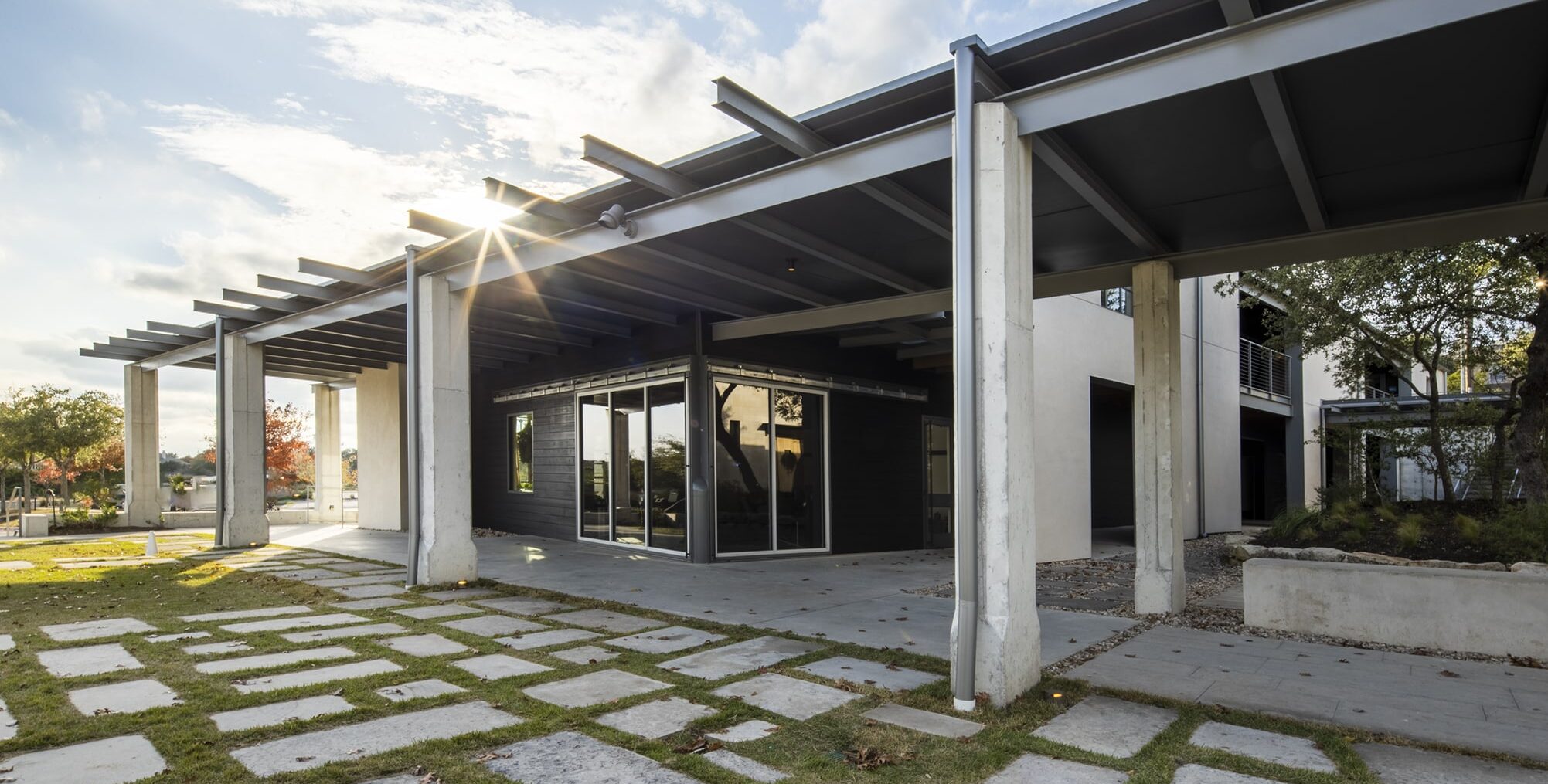 A contemporary building with tall concrete columns, exposed steel beams, and glass walls. A walkway made of rectangular stone slabs set in grass leads to the entrance. Sunlight shines through the roof structure, and trees and landscaped areas surround the building.