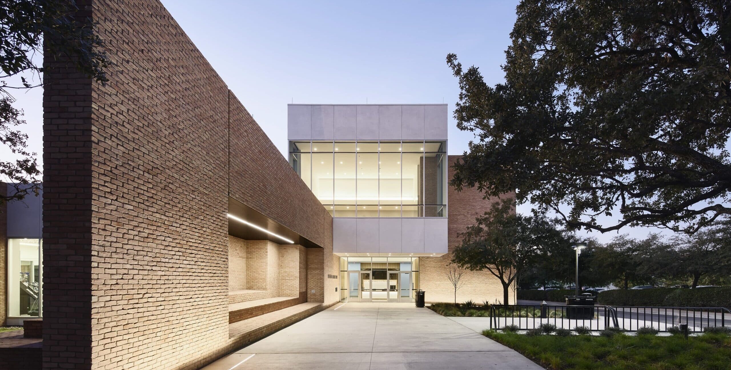 A modern building with brick walls and large glass windows, illuminated at twilight. The entrance is framed by tall brick structures, and exterior lighting highlights the walkway. Trees and a cool evening sky appear in the background.