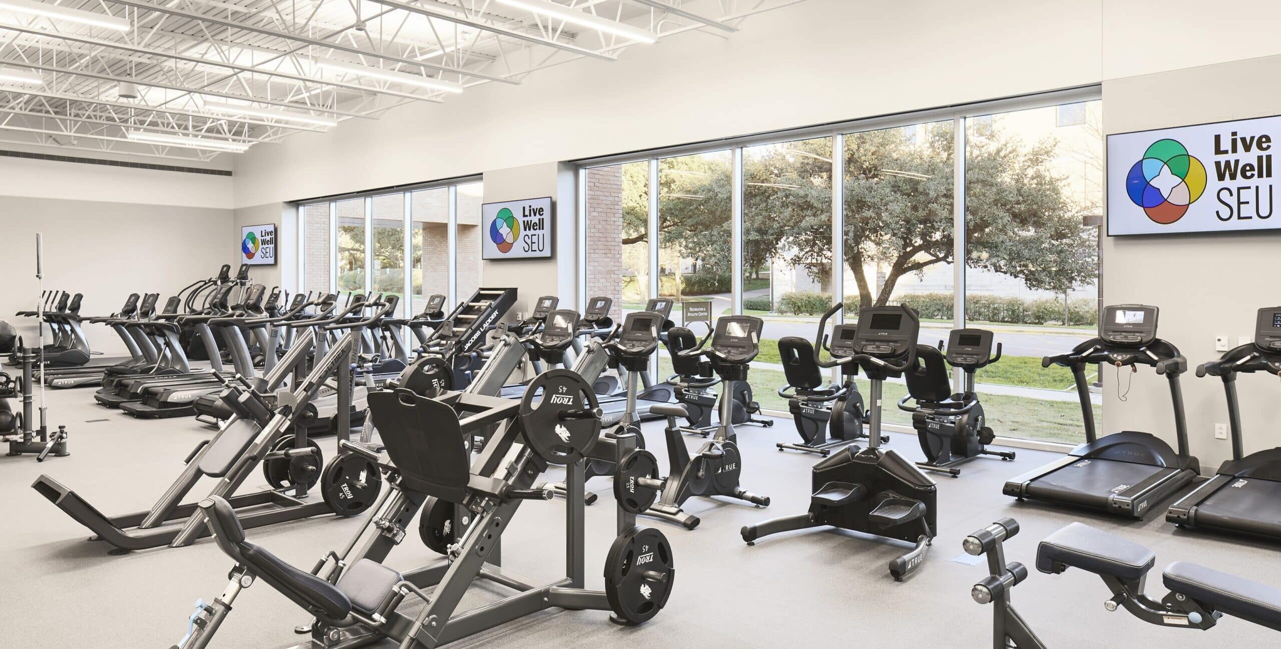 A bright fitness center filled with treadmills, stationary bikes, ellipticals, and weight machines. Large windows show trees outside. Digital screens on the wall display the text “Live Well SEU.” The ceiling has exposed beams and bright lighting.