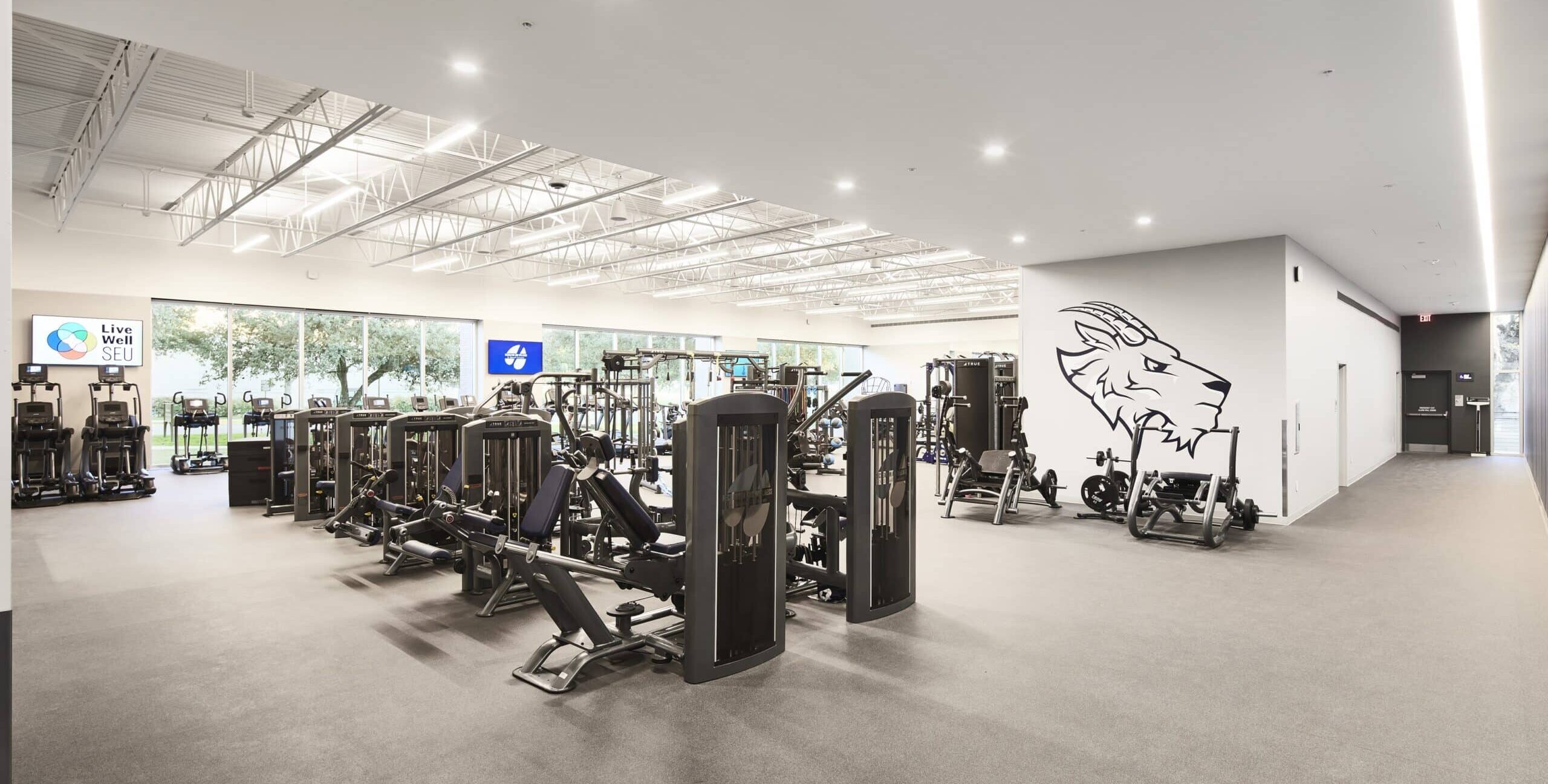 A spacious gym with high ceilings, exposed beams, and bright lighting. The room contains strength machines, cable stations, and cardio equipment. Large windows reveal greenery outside. A stylized goat‑head graphic is displayed on the right wall, and a screen on the left shows “Live Well SEU.”