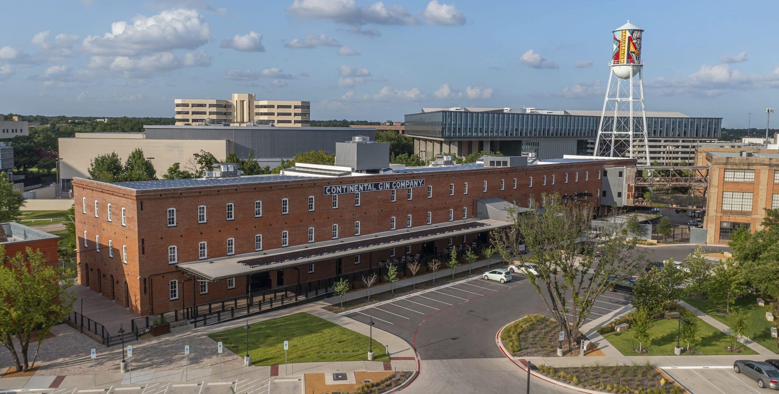 A large brick industrial building with the words “CONTINENTAL GIN COMPANY” on its side. The scene includes a covered loading dock, rows of windows, landscaped trees, a parking lot, and a tall white water tower with colorful artwork.
