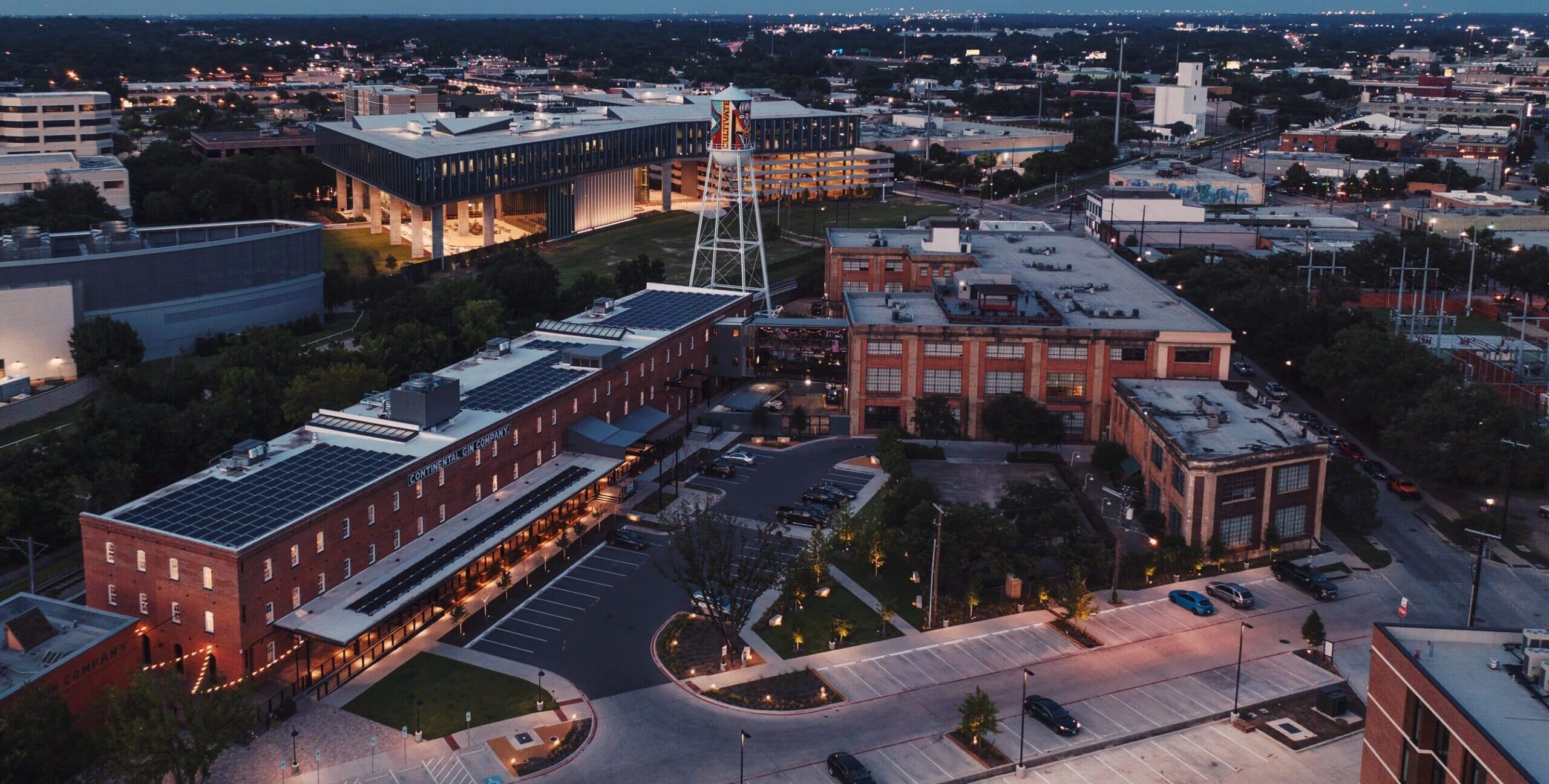 An aerial view of a long red‑brick industrial building with solar panels and the words “CONTINENTAL GIN COMPANY” on its side. Nearby brick structures, a white water tower with artwork, parking lots, trees, and a modern illuminated building appear within a dusk cityscape.