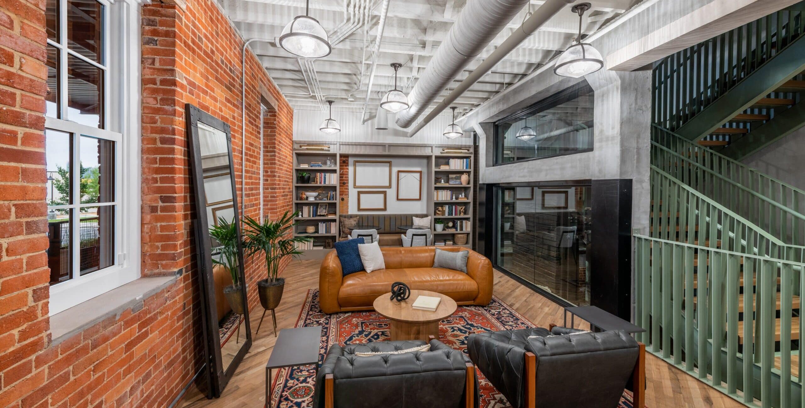 A modern lounge with exposed brick walls, ceiling beams, and ductwork. The seating area includes a tan leather sofa, two black tufted armchairs, and a round wooden coffee table on a patterned rug. Built‑in bookshelves, plants, and a tall mirror decorate the space, and a green staircase rises on the right.