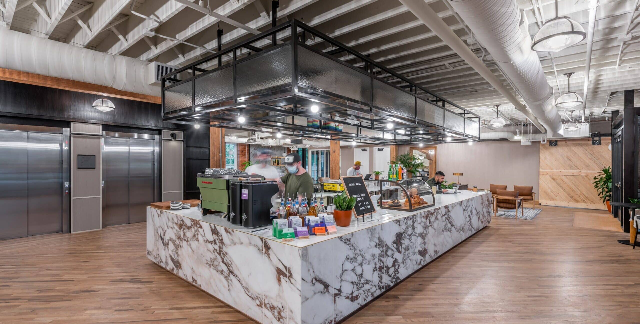 A café counter with a marble-patterned front, a green espresso machine, pastries in a display case, and two people working behind the counter. The space has wooden floors, exposed white ceiling beams, large ducts, pendant lights, and stainless steel elevator doors in the background.