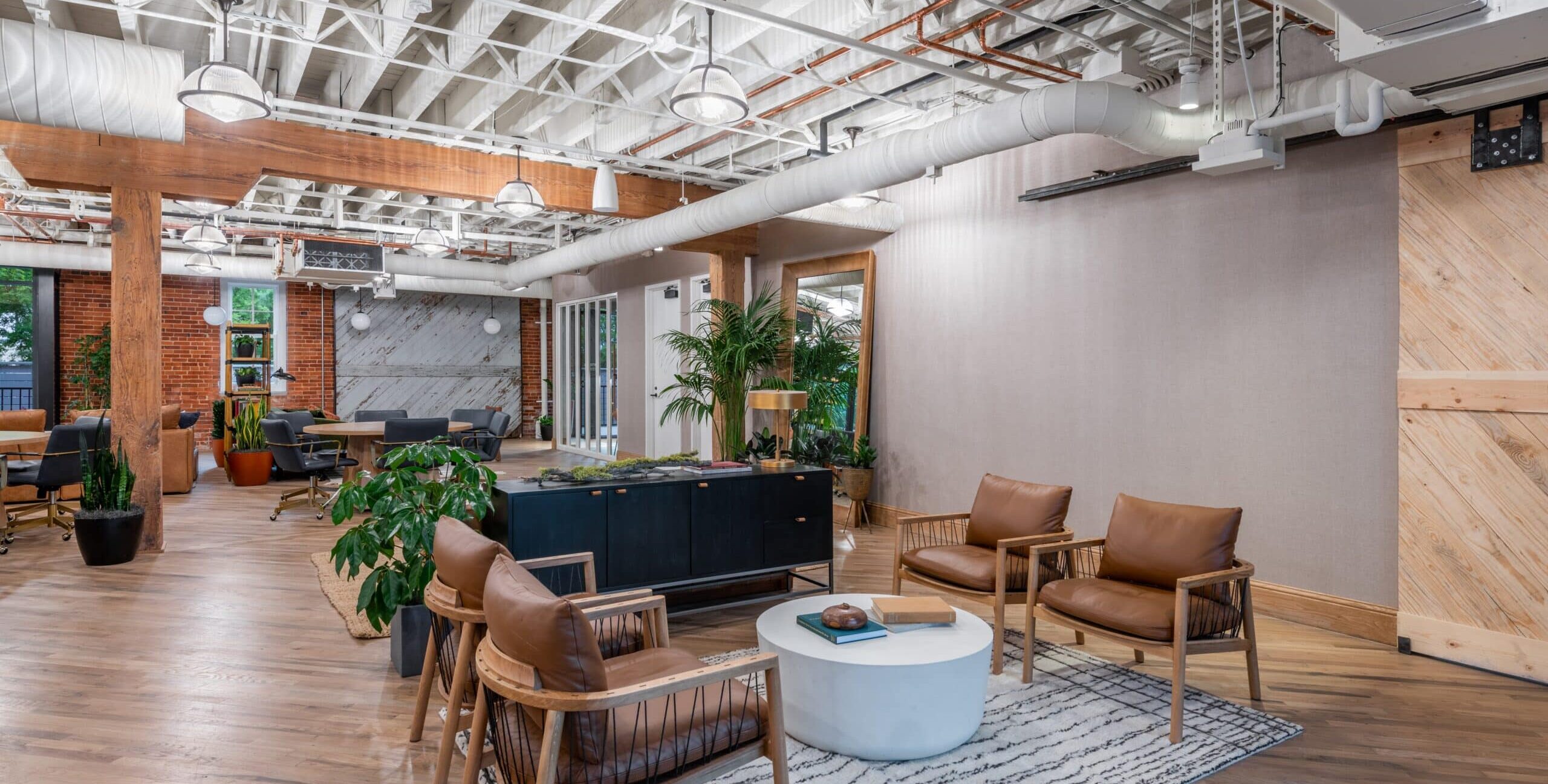 A modern open office with exposed white ceiling beams, ductwork, and wooden support columns. Four wooden-framed chairs with brown leather cushions surround a round white coffee table on a patterned rug. Plants, a black cabinet, and additional seating areas fill the background.