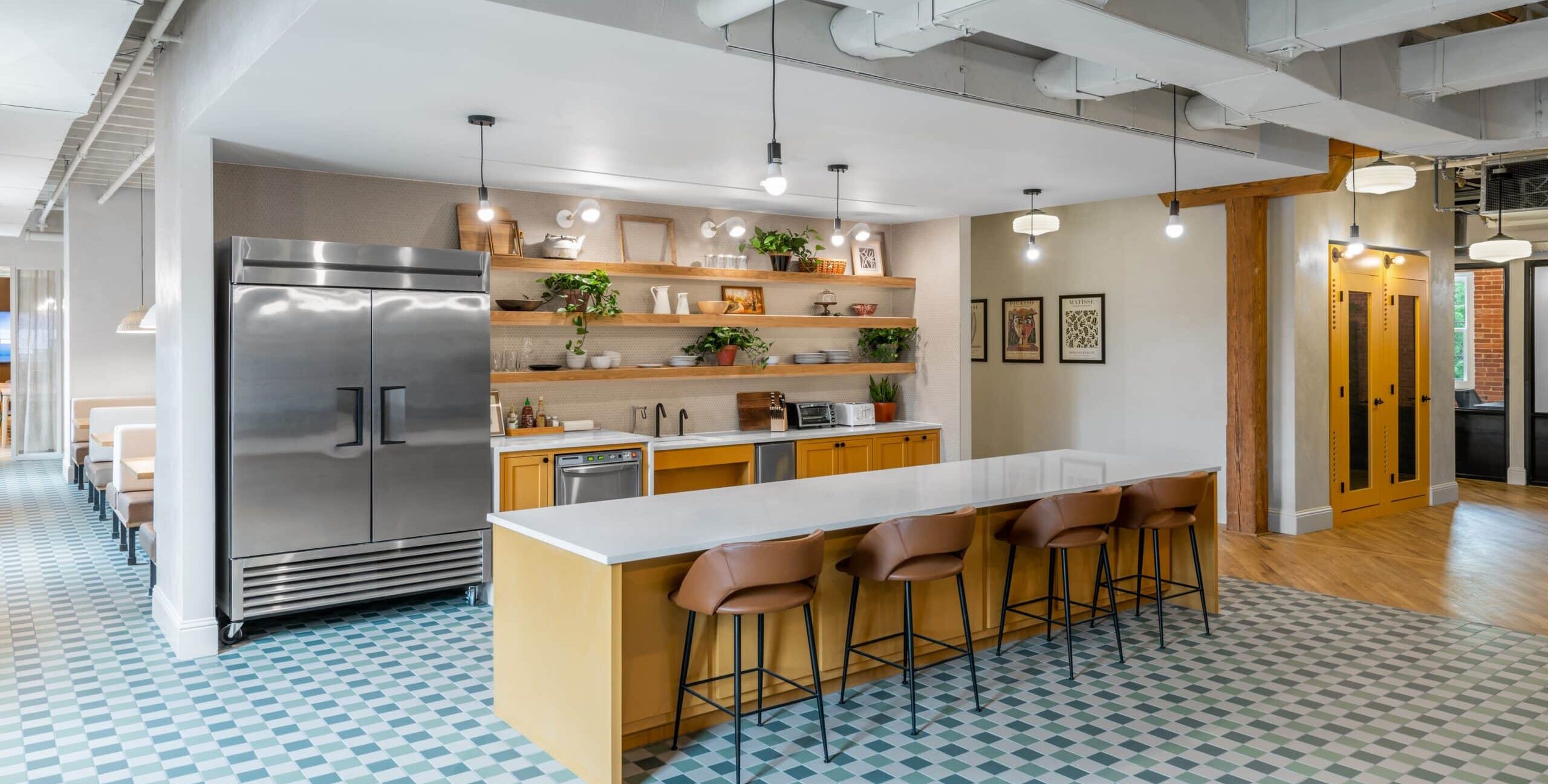 A contemporary shared kitchen with a stainless steel double-door refrigerator, open wooden shelves, yellow lower cabinets, and a long island with four brown bar stools. The floor has blue‑and‑white checkered tiles, and the ceiling features exposed ducts and hanging lights.