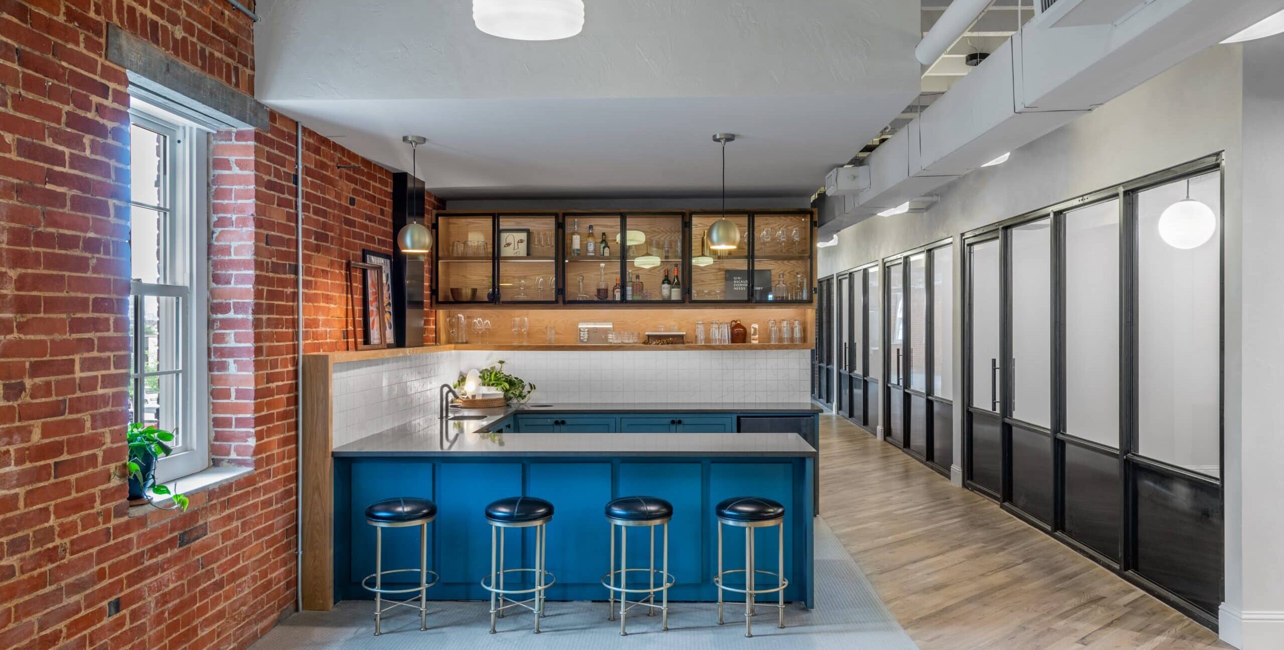 A modern kitchen with exposed red brick walls, a large window, and a blue island with four black cushioned bar stools. Wooden shelves and glass‑front cabinets hold bottles and glassware. Pendant lights hang above the island, and a hallway with glass office doors extends to the right.