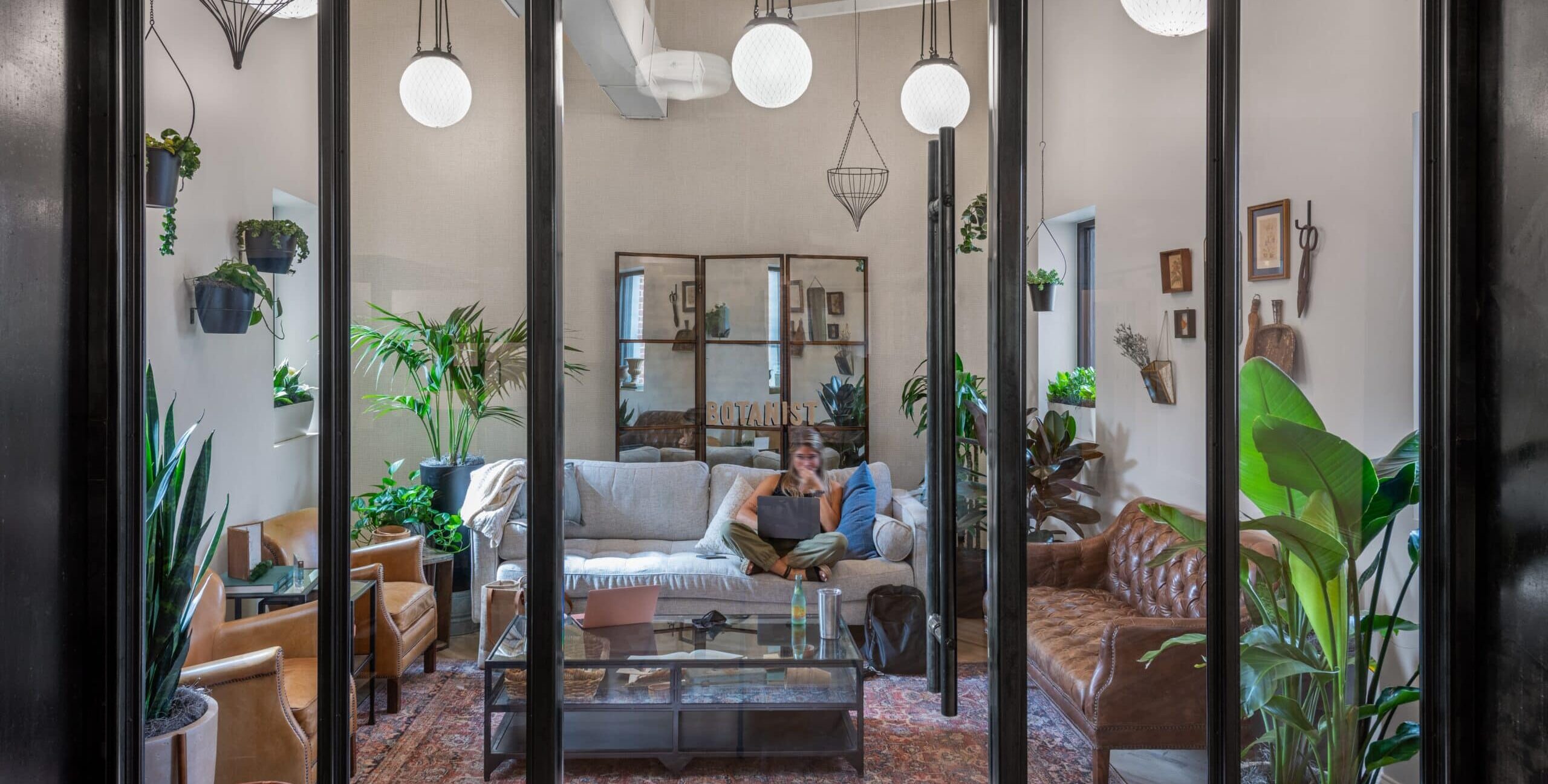 A cozy lounge viewed through a glass wall with black metal framing. Inside are a gray sofa, a brown leather sofa, tan armchairs, and a glass coffee table on a patterned rug. Numerous plants fill the room, and “BOTANIST” appears on a mirrored back wall.