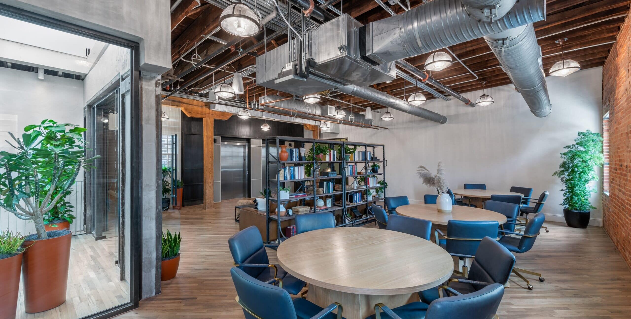A modern office with round wooden tables surrounded by blue upholstered chairs on casters. The space features exposed wooden beams, metal ductwork, industrial lighting, and a large bookshelf filled with books and plants. Stainless steel elevator doors and a seating area appear in the background.