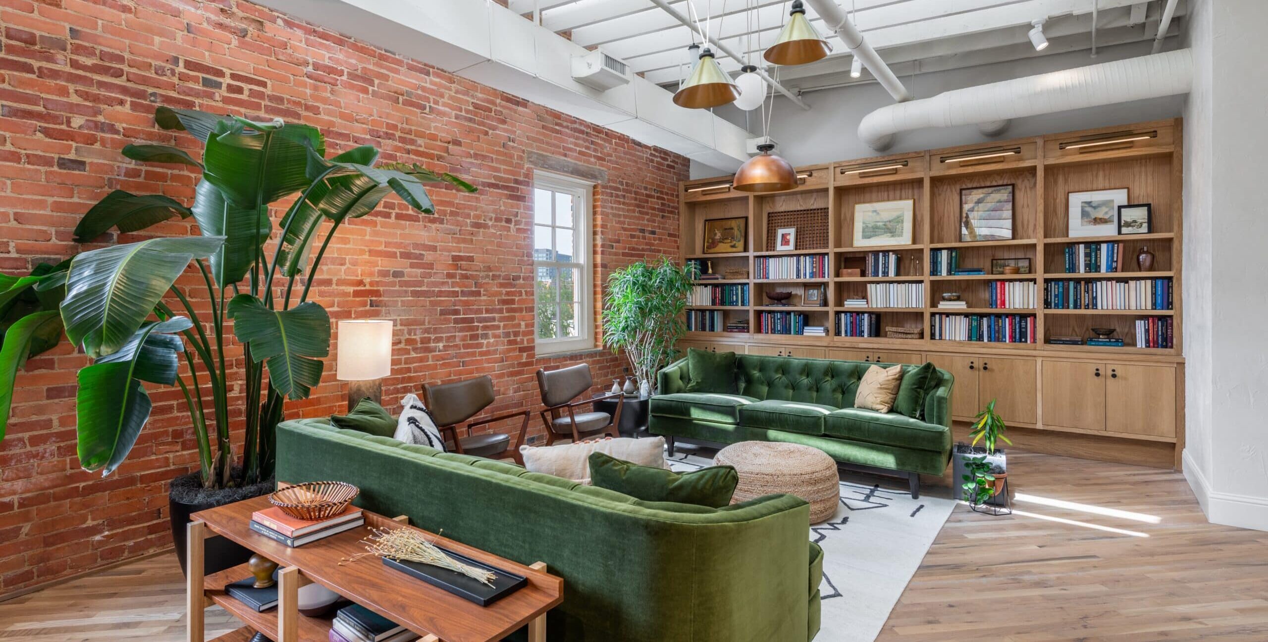 A stylish loft living room with exposed brick walls, white ceiling beams, and large windows. Two green velvet sofas face a woven ottoman on a light rug, with brown leather chairs nearby. Built‑in bookshelves and large potted plants add warmth and color.