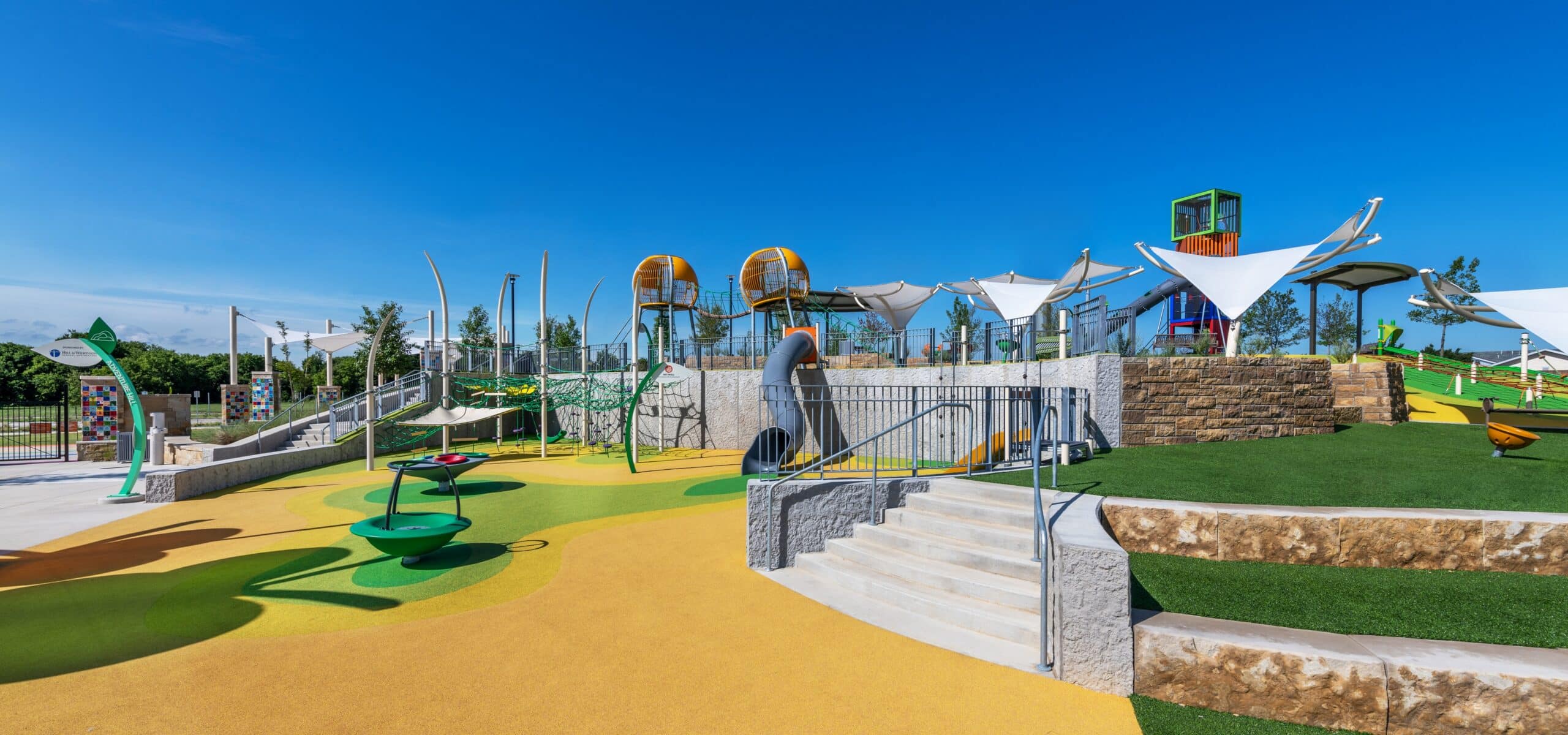 A modern playground featuring elevated platforms connected by bridges, enclosed tube slides, climbing nets, stairs, ramps, and accessible pathways. The ground is covered with yellow and green rubberized safety surfacing, and white shade canopies provide sun protection.