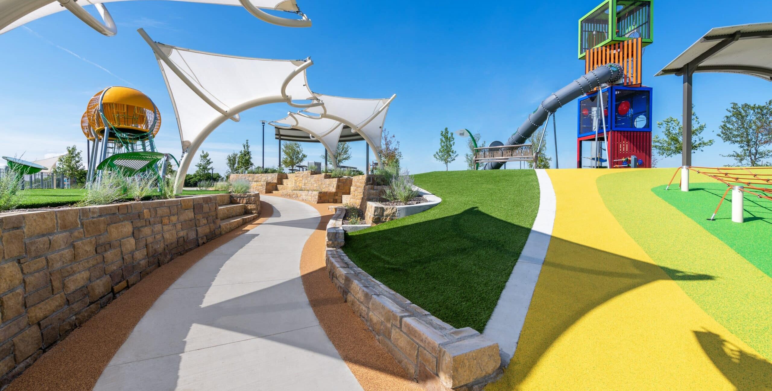 A contemporary outdoor playground with a winding concrete path, stone retaining walls, landscaped plants, a large yellow spherical climbing structure, and a tall multicolored tower with an enclosed tube slide. White shade canopies line the walkway, and the play area sits on green and yellow safety surfacing under a clear blue sky.