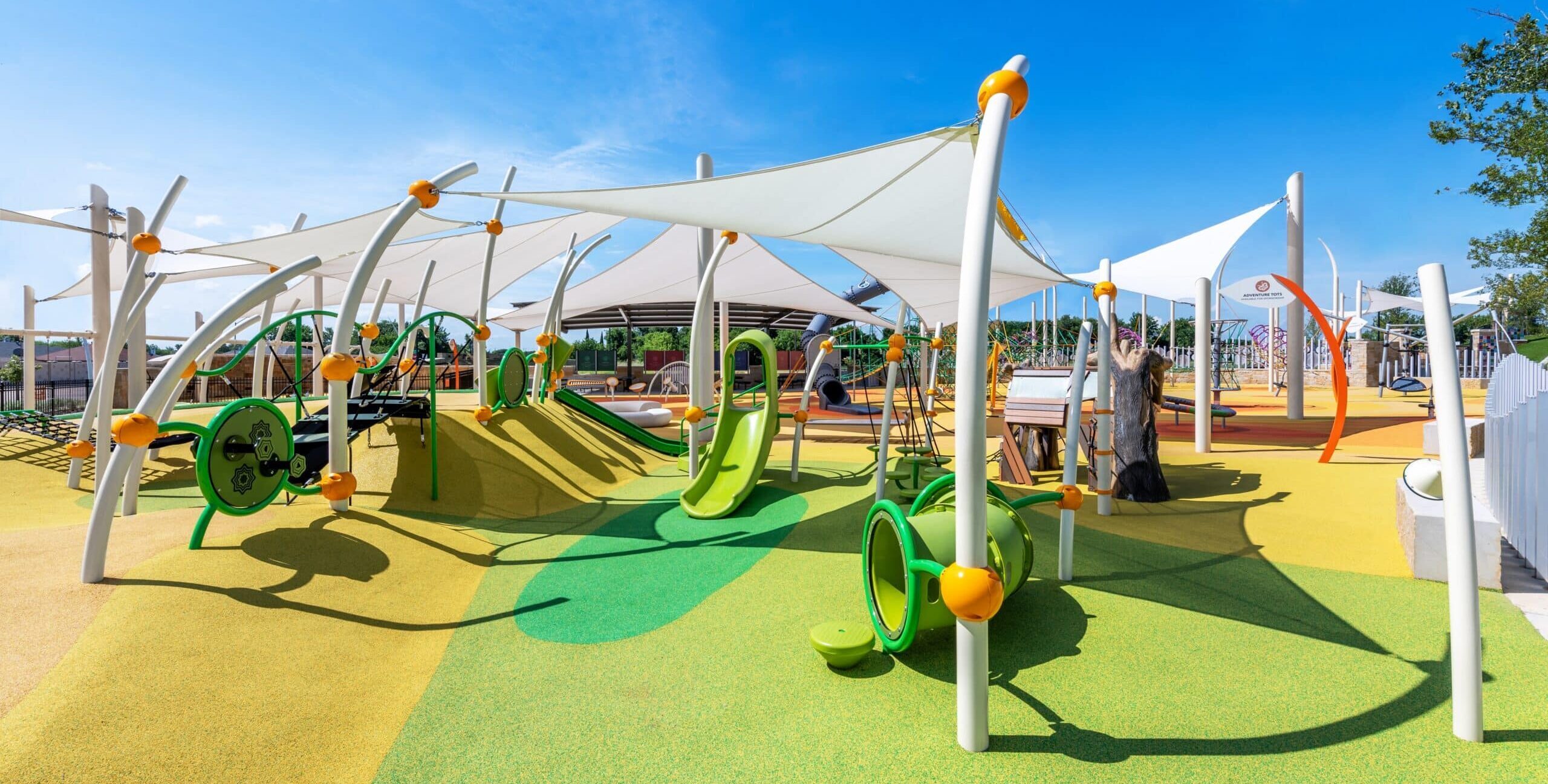 A vibrant playground with green, yellow, and orange play structures including slides, climbing frames, tunnels, and interactive panels. The ground is covered with soft green and yellow rubber surfacing, and large white shade sails are supported by curved poles overhead.