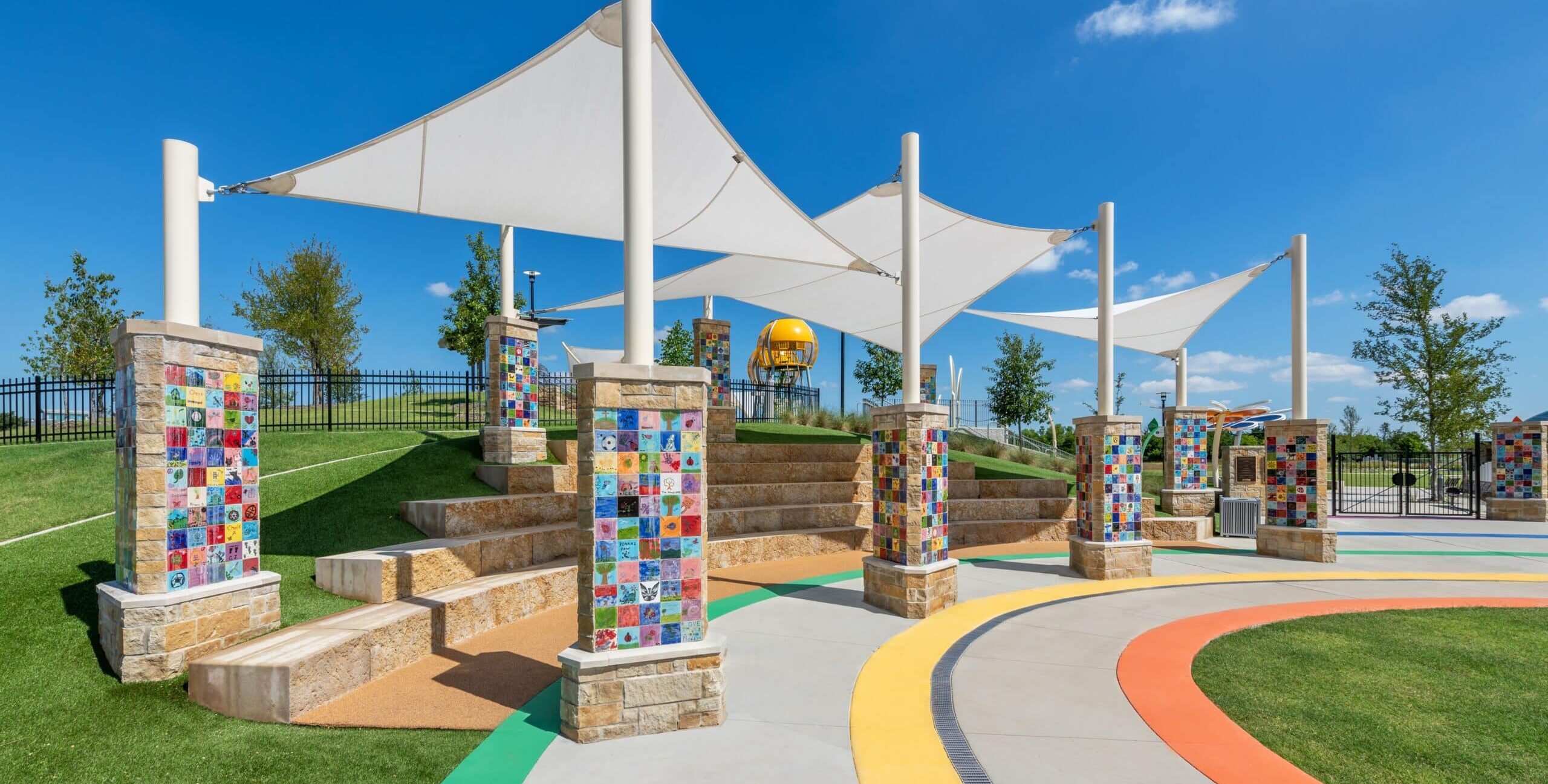 A semicircular outdoor amphitheater with tiered stone seating, large white fabric shade canopies supported by tall poles, and stone pillars decorated with colorful tile artwork. Curved pathways in bright colors run through the grassy area, with playground elements visible in the background.