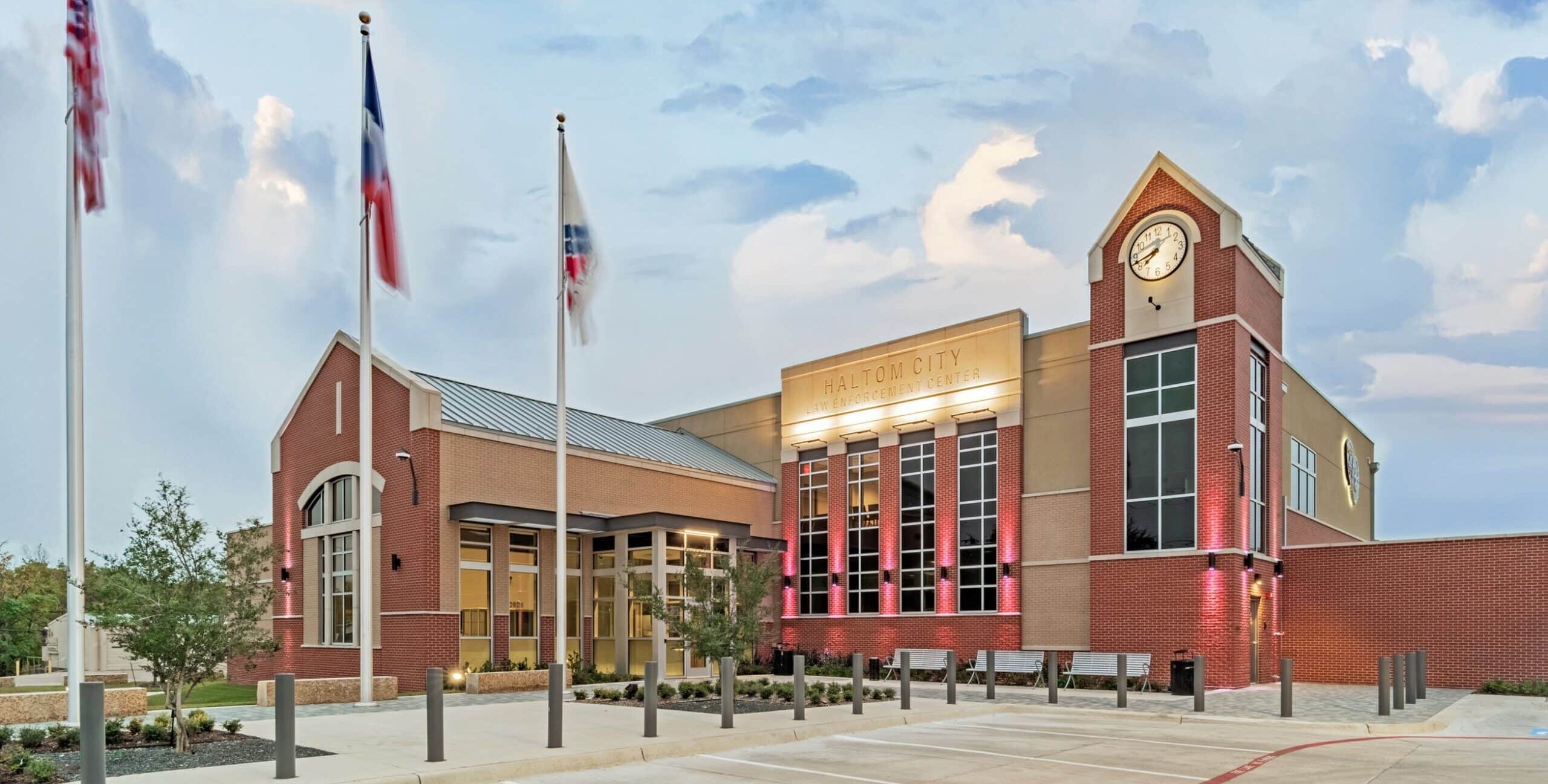 A modern brick and glass civic building with a clock tower, three flagpoles, accent lighting, and landscaped greenery in front of a paved parking area under a partly cloudy evening sky.