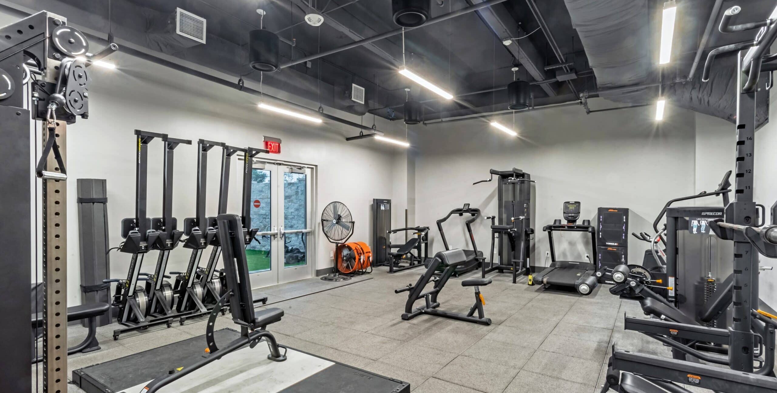 A gym with rowing machines, weight benches, strength‑training machines, gray padded flooring, bright overhead lighting, exposed ductwork, and a double glass exit door.