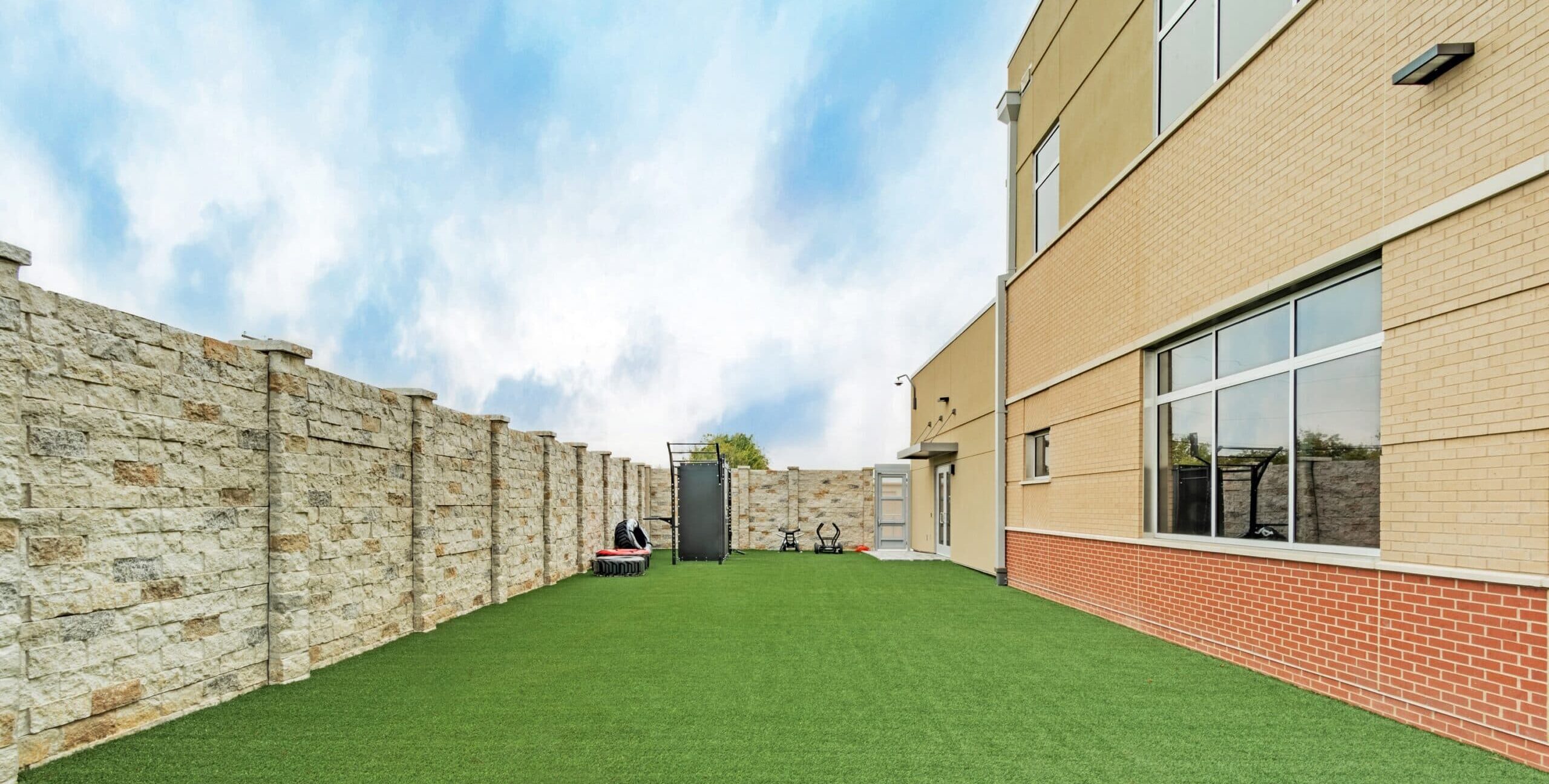 An enclosed outdoor training space with artificial turf, stone walls, and exercise equipment including a trampoline, sled, and stationary bikes beside a modern brick building.