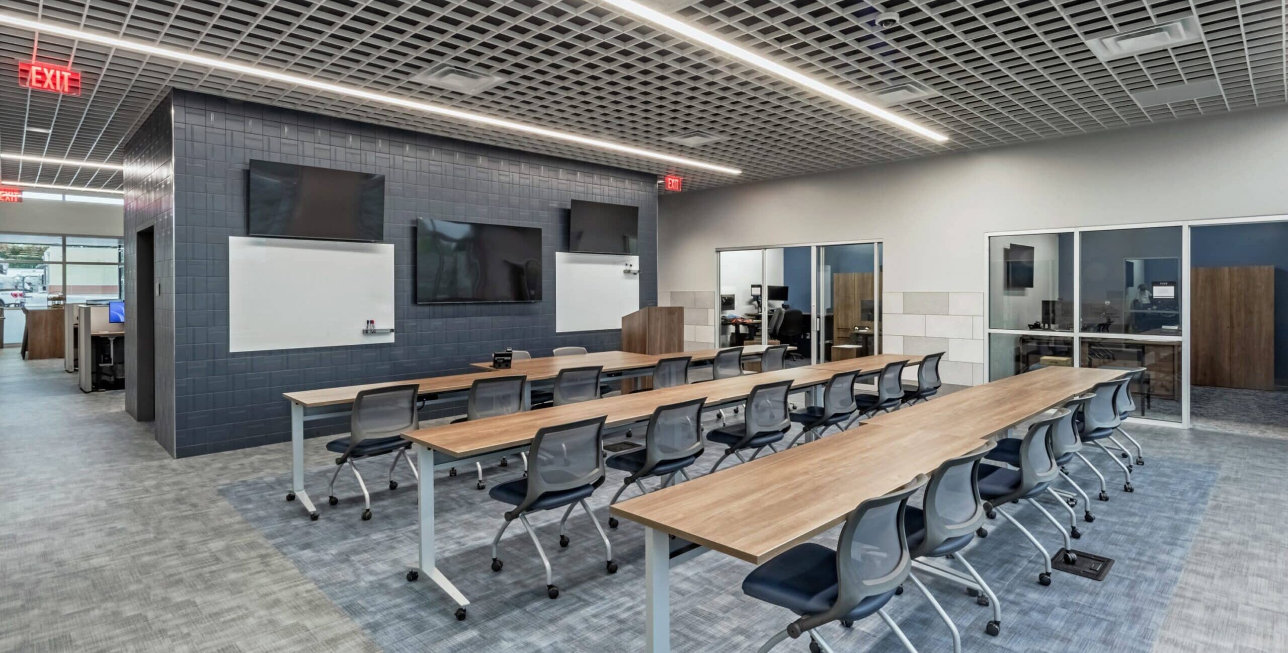 A modern training room with long wooden tables, gray rolling chairs, three large monitors, two whiteboards, a lectern, and glass walls revealing adjacent office spaces.