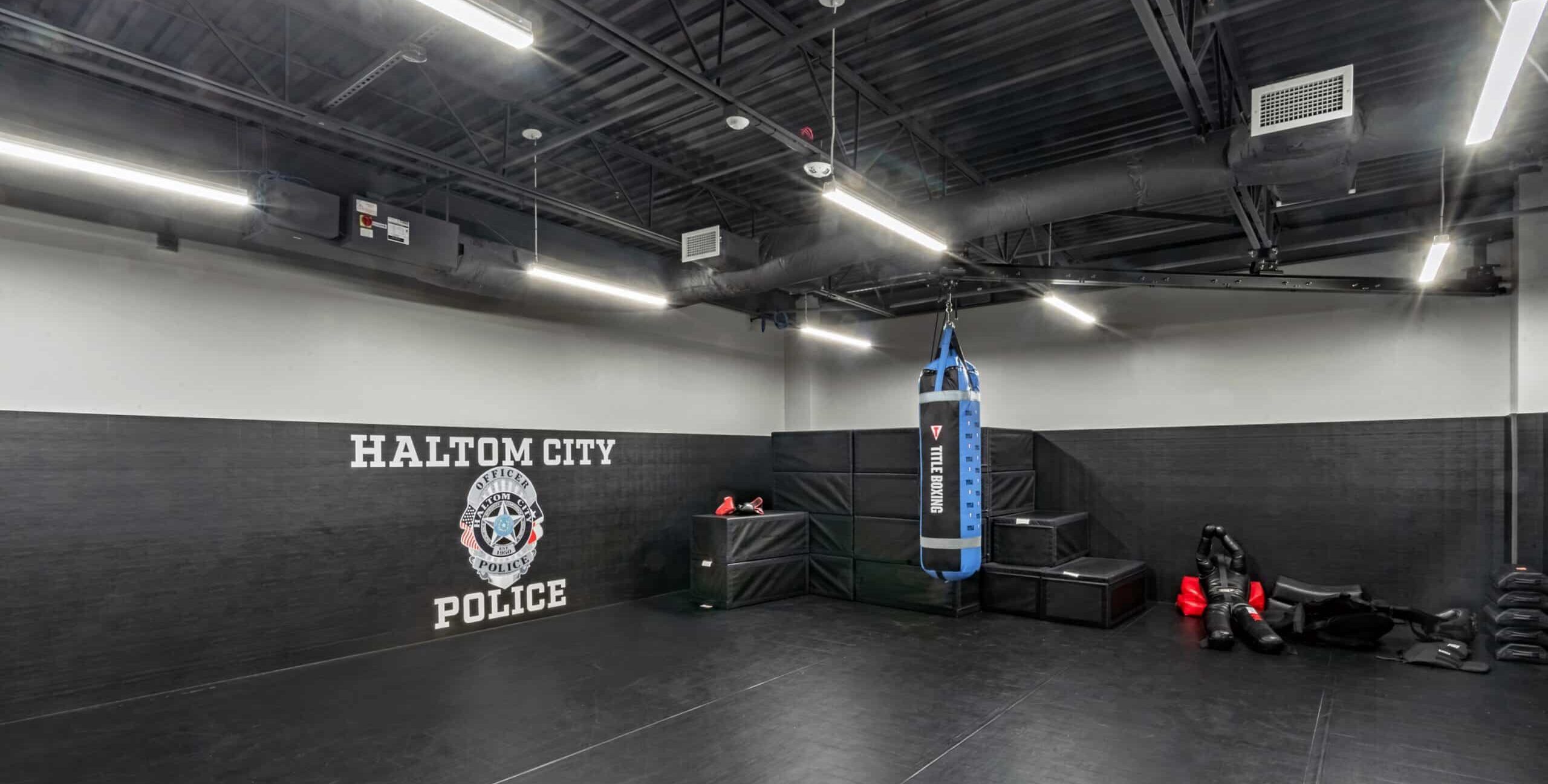 A padded training room with black flooring and walls, a printed police logo, a hanging punching bag, stacked padded boxes, and red and black training dummies under exposed ceiling beams.